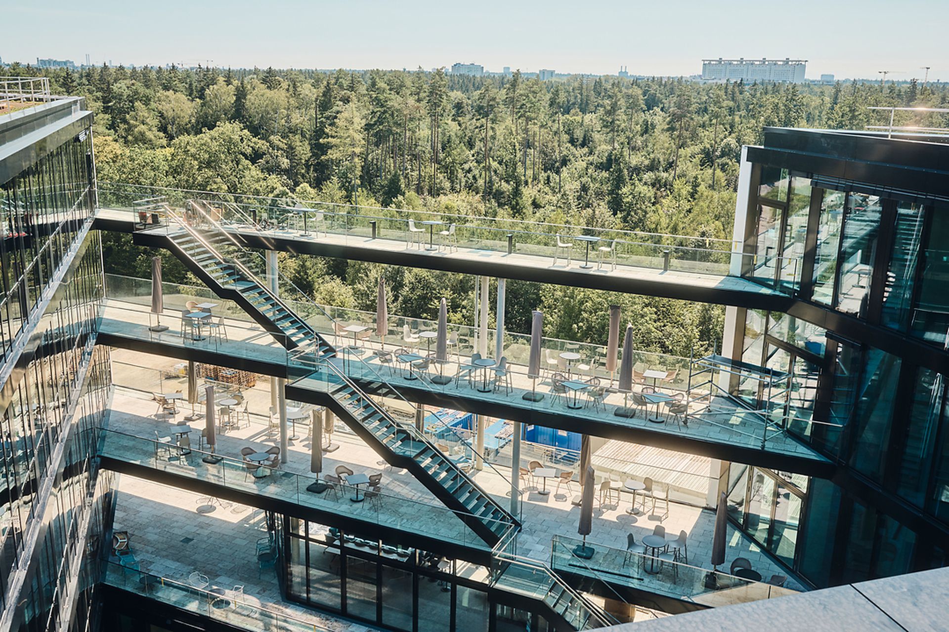 Modernes mehrstöckiges Gebäude mit Glaswänden, Außenterrassen und Treppen, umgeben von üppigem grünen Wald unter klarem Himmel.