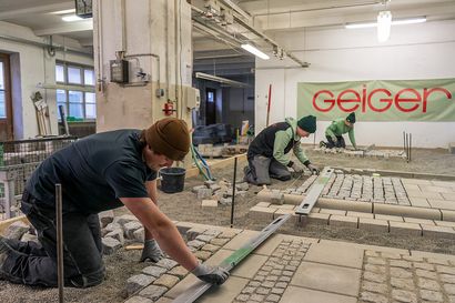 Drei Auszubildende von Geiger führen Pflasterarbeiten im Rahmen der Winterausbildung durch. Sie verlegen Kopfsteinpflaster und Betonplatten, während Werkzeuge und Richtschnüre zur genauen Ausrichtung genutzt werden. Im Hintergrund hängt ein Geiger-Banner.