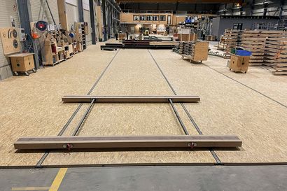 Blick entlang der Fertigungslinie bei Geiger Holzsystembau. Holzplattformen und Schienen durchziehen den Raum, während im Hintergrund Arbeitsbereiche und Regale für Materialien sichtbar sind.