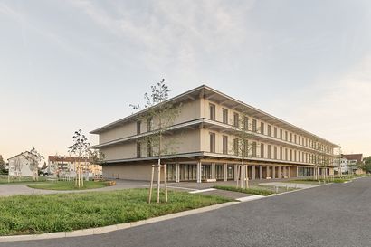 Ein zweigeschossiger Schulneubau aus Holz im Fokus mit Grünanlagen im Vordergrund.