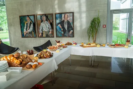 Ein Buffettisch mit einer Auswahl an Broten, Käsesorten, Fleischwaren und Snacks in einem Raum. An der Wand hängen drei Porträts, und neben dem Fenster befindet sich eine große Pflanze