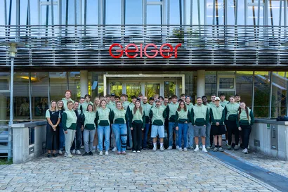 Eine Gruppe von Leuten in grünen Hemden steht vor einem Gebäude mit Glasfront und einem roten "Geiger"-Schild.