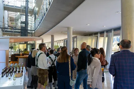 Eine Gruppe von Menschen mischt sich in einer modernen Lobby mit großen Fenstern und einer Treppe, einige tragen traditionelle Kleidung; Getränke stehen auf einem Tisch.