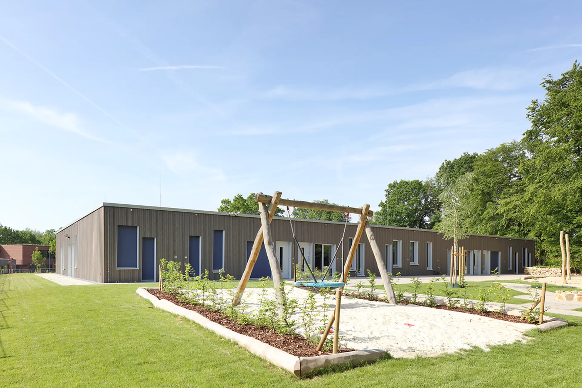 Modernes, eingeschossiges Gebäude mit Holzfassade, umgeben von einer gepflegten Grünfläche. Im Außenbereich befinden sich eine Schaukelanlage und ein Sandspielbereich. Die freundliche Anlage liegt unter einem klaren blauen Himmel und bietet eine einladende Umgebung zum Spielen und Verweilen.