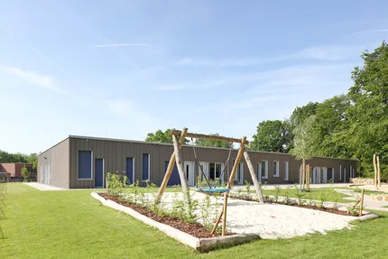 Modernes, eingeschossiges Gebäude mit Holzfassade, umgeben von einer gepflegten Grünfläche. Im Außenbereich befinden sich eine Schaukelanlage und ein Sandspielbereich. Die freundliche Anlage liegt unter einem klaren blauen Himmel und bietet eine einladende Umgebung zum Spielen und Verweilen.