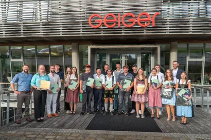 Eine Gruppe von Menschen hält Zertifikate und Mappen in den Händen und steht vor einem Gebäude mit einem „Geiger“-Schild.