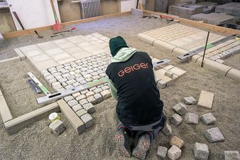 Ein Auszubildender von Geiger arbeitet kniend an einer Pflasterfläche, die aus Betonplatten und Kopfsteinpflaster besteht. Vor ihm liegen Werkzeuge wie eine Wasserwaage und eine Richtschnur. Der Geiger-Schriftzug ist auf der Arbeitskleidung zu sehen.