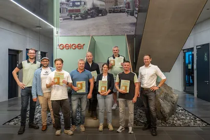 Eine Gruppe von neun lächelnden Personen hält Urkunden in den Händen und posiert in einer modernen Lobby vor einem Geiger-Schriftzug sowie einem historischen Foto eines Lastwagens an der Wand.  Geben Sie Ihr Feedback zu BizChat ab.