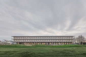 Ein zweigeschossiger Schulneubau aus Holz im Fokus mit Grünanlagen im Vordergrund.