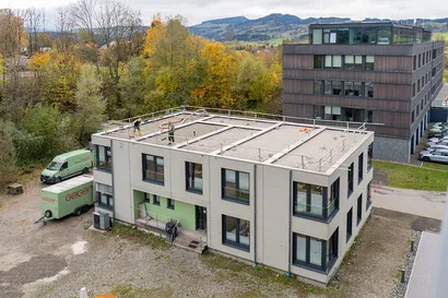 Zwei Arbeiter auf dem Flachdach eines grauen, zweistöckigen Gebäudes, umgeben von Bäumen und Autos, mit einem weiteren Gebäude im Hintergrund.