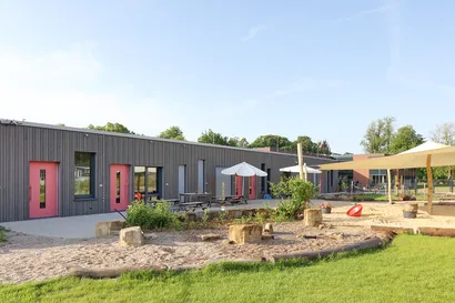 Modernes Gebäude mit roten Türen und großzügigen Fensterflächen. Im Außenbereich befinden sich ein Spielplatz mit Sonnenschirmen, Picknicktischen, Sandspielfläche und viel Grün. Die Anlage liegt unter einem klaren blauen Himmel und bietet eine freundliche, einladende Umgebung.