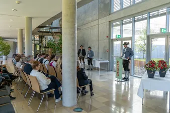 Ein Mann in traditioneller Kleidung spricht an einem Podium zu einem Publikum, das in Reihen in einem modernen Gebäude mit großen Fenstern und Topfpflanzen sitzt.