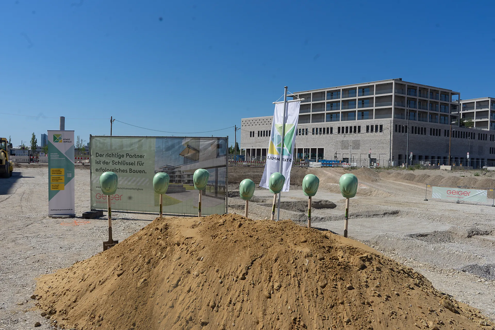 Baustelle mit aufgestellten Spaten vor einem Erdhaufen, umgeben von Bannern und Flaggen. Im Hintergrund befindet sich ein großes Gebäude.