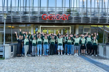 Eine Gruppe von Leuten in grünen Hemden steht vor einem Gebäude mit Glasfront und einem roten "Geiger"-Schild und jubeln..