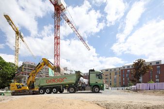 Ein Bagger lädt einem LKW von Geiger Material auf. Im Hintergrund sieht man eine Baustelle.