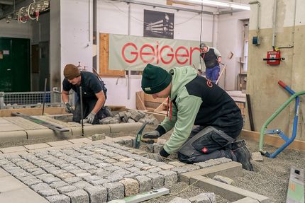 Zwei Auszubildende von Geiger bei der Verlegung von Kopfsteinpflaster. Eine Person nutzt einen Hammer, während die andere Pflastersteine aus einem Eimer vorbereitet. Die Szene zeigt Werkzeuge und Baumaterialien in einem Ausbildungsraum.