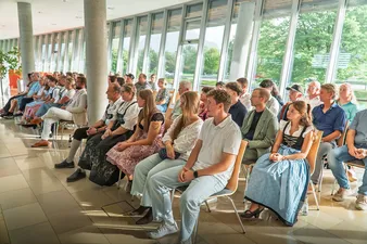 Eine vielfältige Gruppe von Menschen sitzt aufmerksam in Reihen. Einige tragen traditionelle Kleidung. Der Raum ist von Tageslicht durchflutet und verfügt über große Fenster mit Blick ins Grüne.