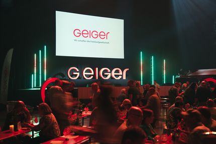 Eine Geiger-Veranstaltung bei schwacher Beleuchtung mit Menschen an Tischen. Im Hintergrund ist auf einer großen Leinwand und in Leuchtbuchstaben „Geiger“ zu sehen.