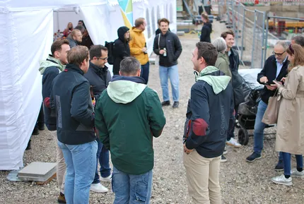 Eine Gruppe von Personen in Jacken unterhält sich bei einer Veranstaltung im Freien in der Nähe eines weißen Zeltes. Der Boden ist mit Kies bedeckt, im Hintergrund ist eine Baustelle zu sehen.
