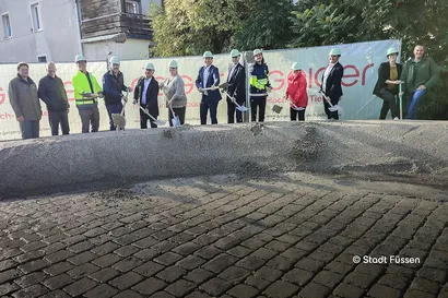 Eine Gruppe von Personen mit Schutzhelmen nimmt an einem Spatenstich teil und schaufelt Erde vor einem Bauschild auf einer Baustelle.