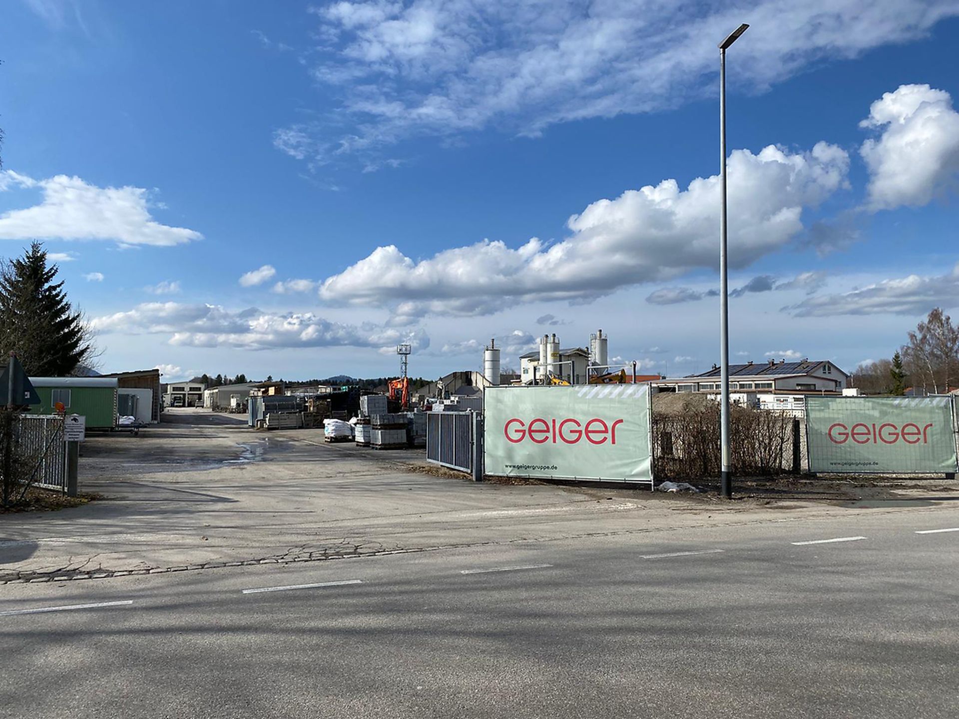 Industriegelände mit eingezäuntem Perimeter, gekennzeichnet mit „Geiger“. Gebäude und Baumaterialien unter blauem Himmel mit vereinzelten Wolken sichtbar.