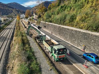 Geiger Baustelle entlang der Kemptner Straße (B308) in Immenstadt. Ein Bagger mit Geiger-Branding lädt Erdreich in einen Lkw, während parallel dazu ein Schienenweg verläuft. Die Baustelle befindet sich an einem Hang mit begrünten Flächen und einer Stützmauer aus Naturstein.