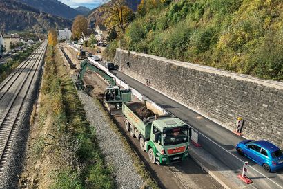 Geiger Baustelle entlang der Kemptner Straße (B308) in Immenstadt. Ein Bagger mit Geiger-Branding lädt Erdreich in einen Lkw, während parallel dazu ein Schienenweg verläuft. Die Baustelle befindet sich an einem Hang mit begrünten Flächen und einer Stützmauer aus Naturstein.
