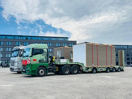 Ein grüner und weißer Lastwagen steht neben einem anderen Lastwagen und transportiert eine große, kastenartige Struktur auf einem Flachbettanhänger in einem Industriegebiet.