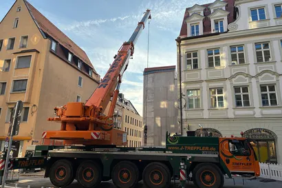 Ein großer orange-grüner mobiler Kran steht zwischen zwei hohen Gebäuden auf einer Stadtstraße und streckt seinen Arm nach oben.