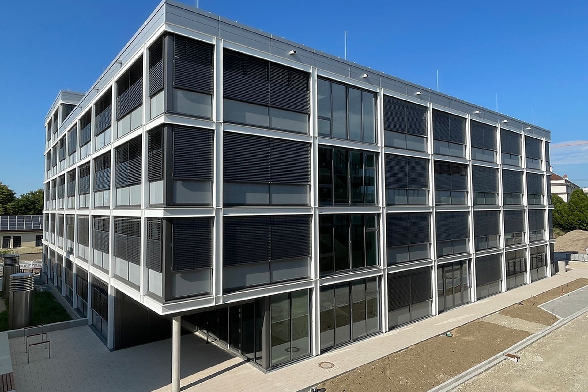 Modernes vierstöckiges Gebäude mit großen Glasfenstern und Metallrahmen unter einem klaren blauen Himmel.