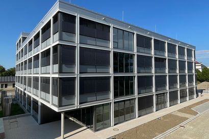 Modernes vierstöckiges Gebäude mit großen Glasfenstern und Metallrahmen unter einem klaren blauen Himmel.