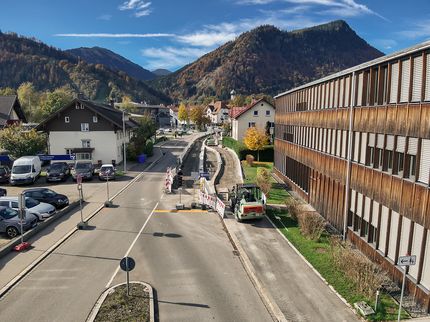 Blick auf die Baustelle in der Kemptner Straße (B308) in Immenstadt. Eine Teilsperrung der Straße zeigt vorbereitende Arbeiten für Infrastrukturmaßnahmen. Man sieht einen Radlader im Geiger-Branding. Die Umgebung umfasst Wohnhäuser, ein modernes Holzgebäude sowie Berge im Hintergrund.