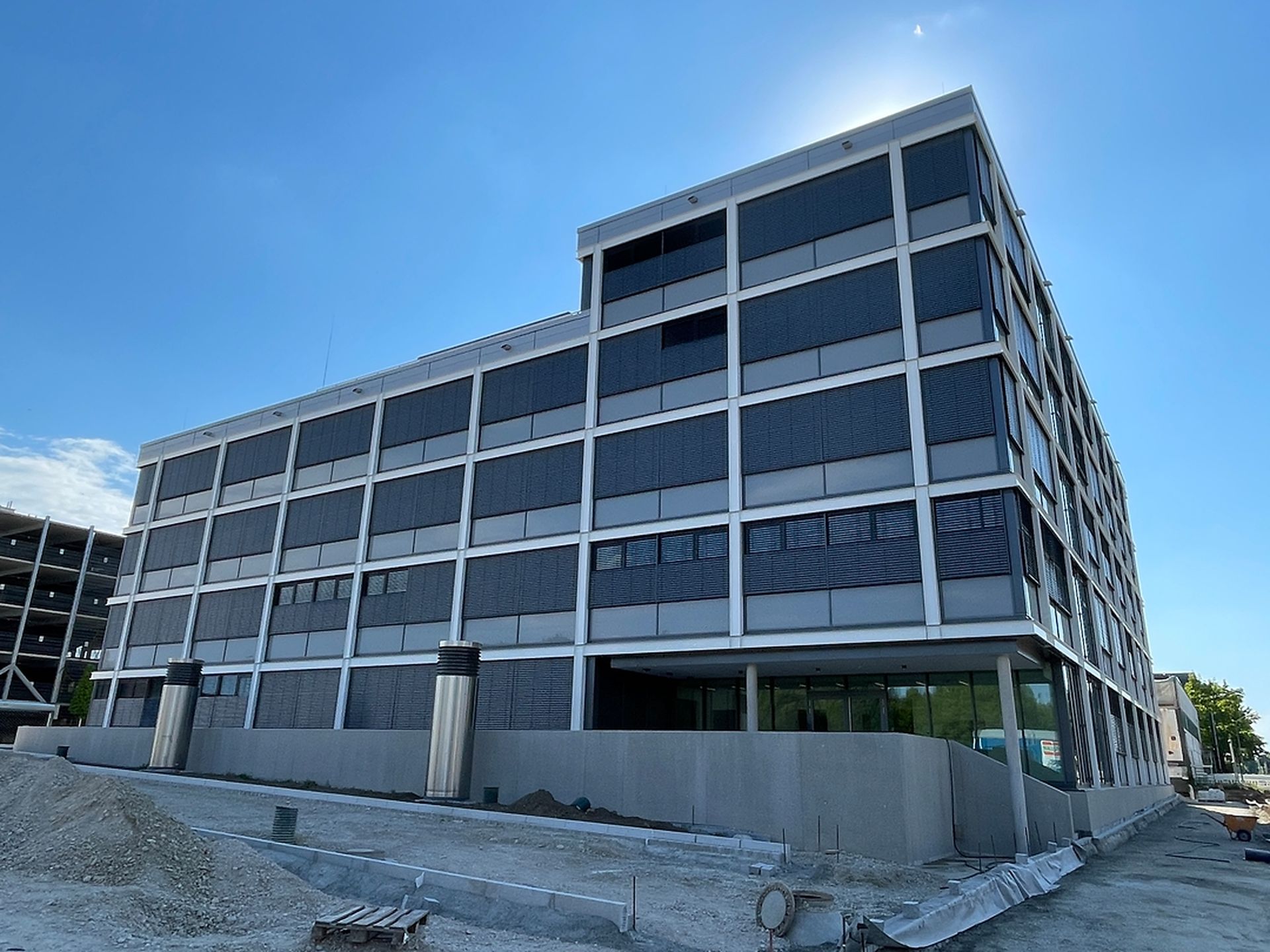 Modernes, mehrstöckiges Gebäude im Bau mit einer gitterartigen Fassade, umgeben von Baumaterialien und einem klaren blauen Himmel darüber.