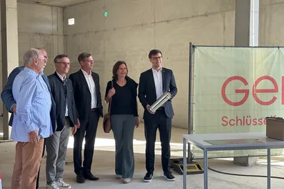 Eine Gruppe von sechs Personen in Geschäftskleidung steht in einem Betongebäude, eine Person hält einen Metallzylinder. Ein Schild und ein Tisch sind neben ihnen zu sehen.