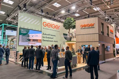 Antwort wird generiert Copilot said:  Personen versammeln sich an einem Messestand mit einem großen Geiger-Schild, der über einen Baum und multimediale Displays verfügt, unter heller industrieller Beleuchtung.
