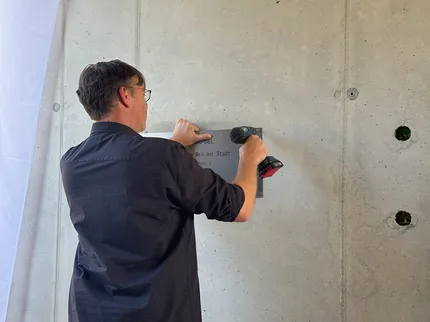 Eine Person in einem schwarzen Hemd benutzt eine Bohrmaschine, um ein Schild an einer Betonwand anzubringen.