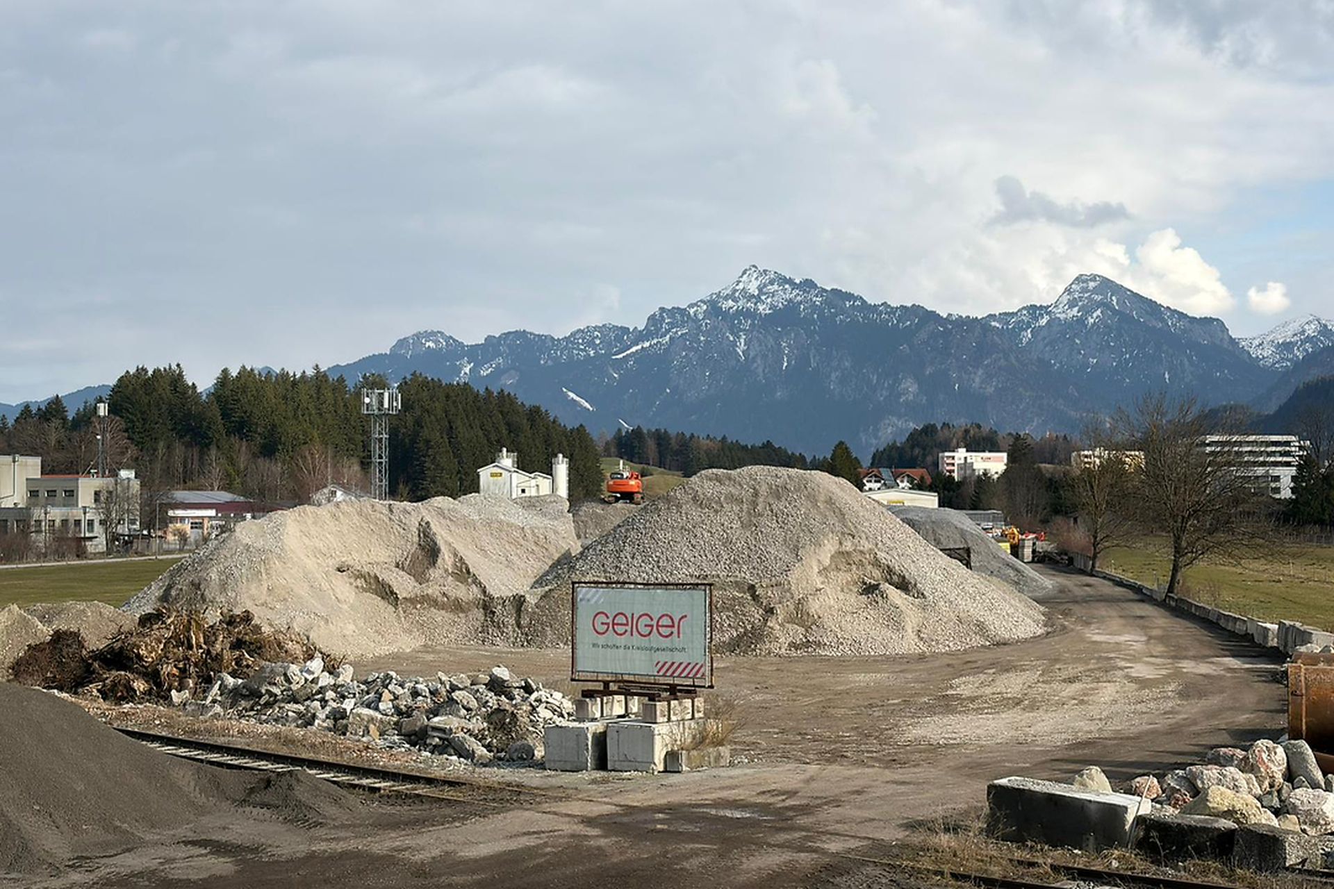 Baustelle mit großen Kieshaufen und Maschinen, Bäumen und schneebedeckten Bergen im Hintergrund unter einem bewölkten Himmel.