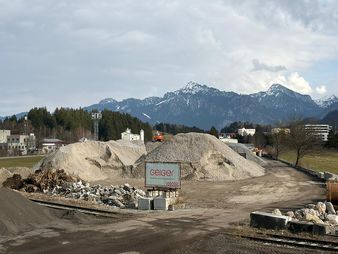 Baustelle mit großen Kieshaufen und Maschinen, Bäumen und schneebedeckten Bergen im Hintergrund unter einem bewölkten Himmel.