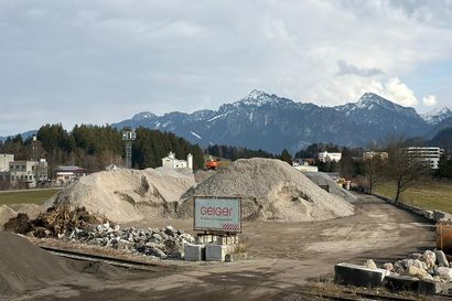 Baustelle mit großen Kieshaufen und Maschinen, Bäumen und schneebedeckten Bergen im Hintergrund unter einem bewölkten Himmel.