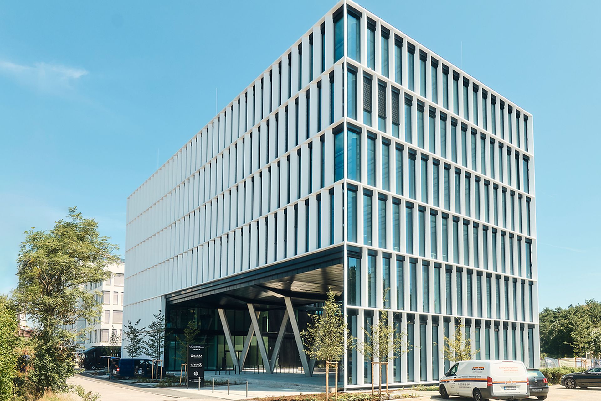 Modernes Bürogebäude mit gitterartiger Fassade, großen Fenstern und überdachtem Eingang. An einem sonnigen Tag parkt in der Nähe ein weißer Lieferwagen.
