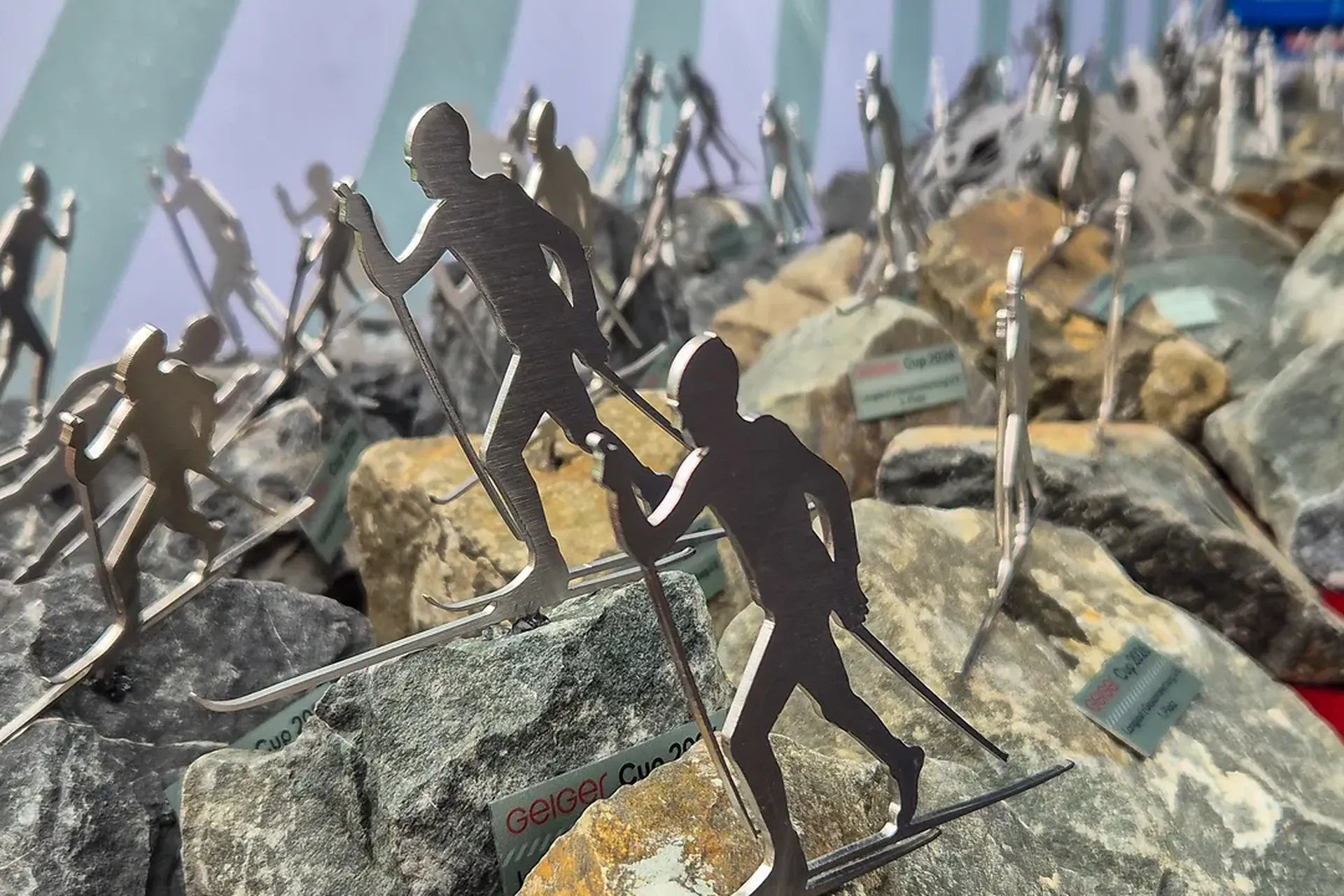 Trophäenanzeige mit Metallskifiguren auf felsigen Sockeln, vor einem Hintergrund mit wirbelnden Streifen und einem Blick auf die Berge.