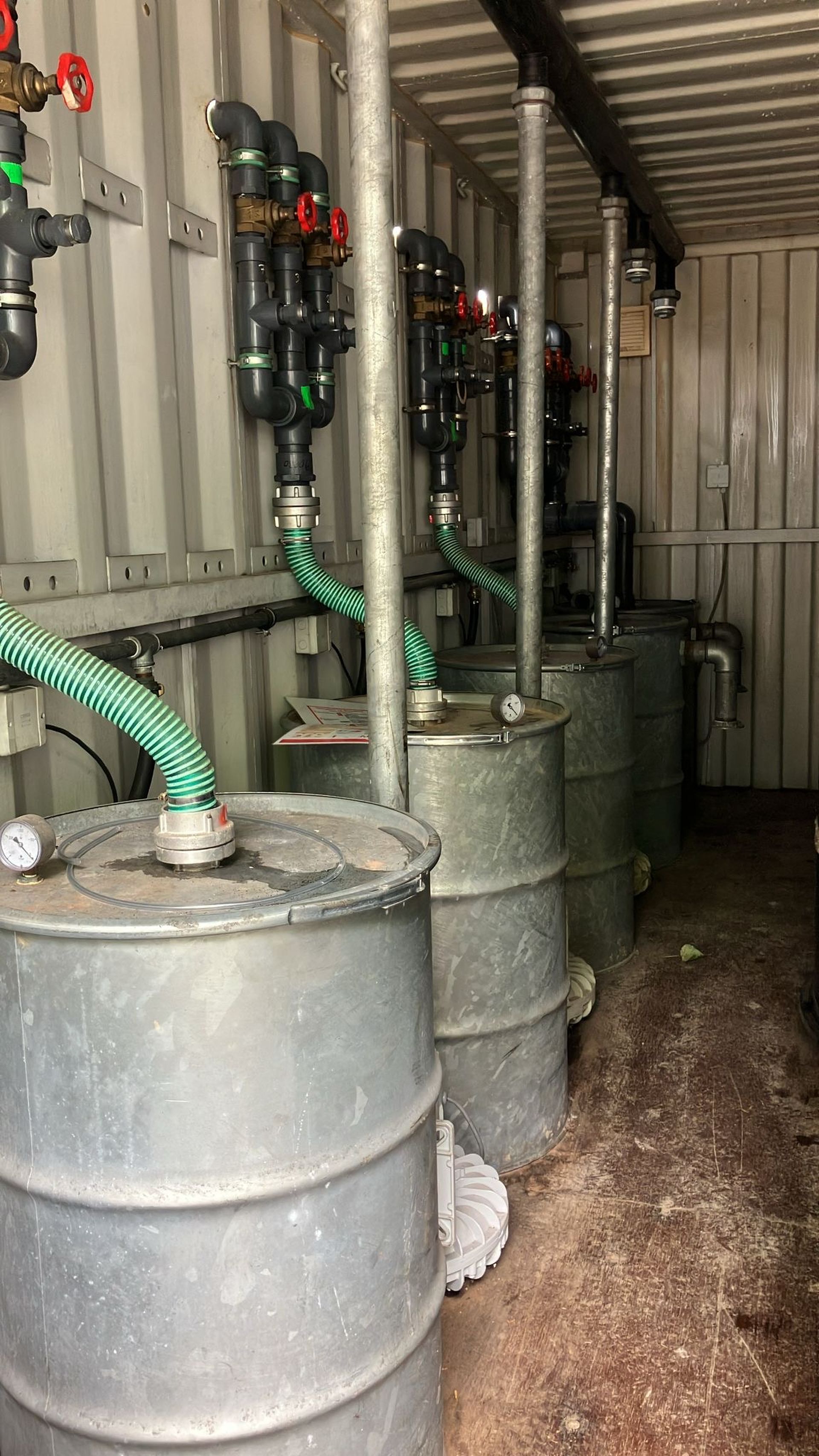 Industrieller Lagerraum mit Metallfässern, die durch grüne Schläuche und Rohre verbunden sind, mit Messgeräten und roten Ventilgriffen an den Wänden.