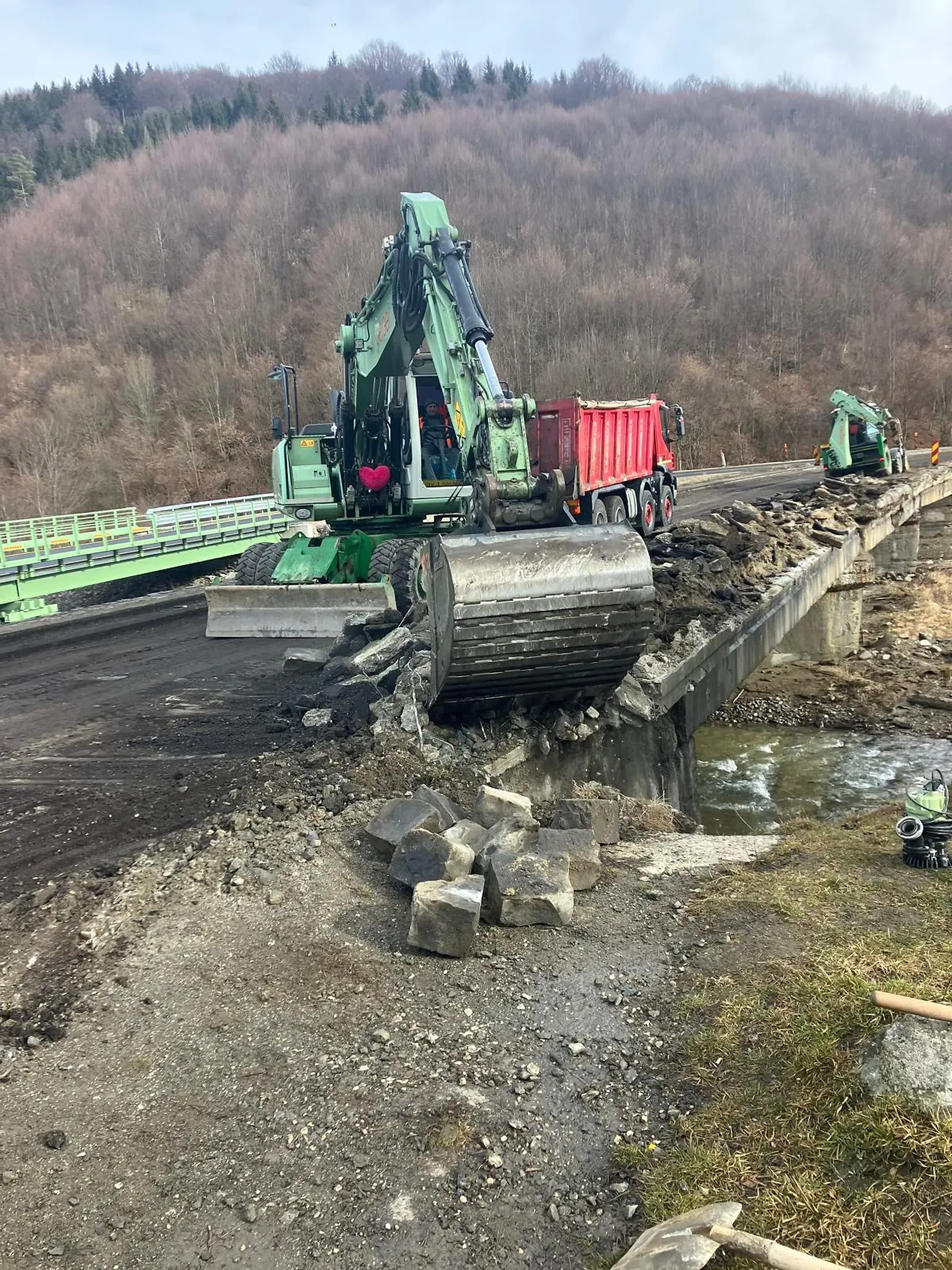 Excavatorul demolează un pod, cu dărâmăturile căzând într-un râu dedesubt. Un camion basculant și un alt excavator sunt în apropiere. Dealuri și pădure în fundal.