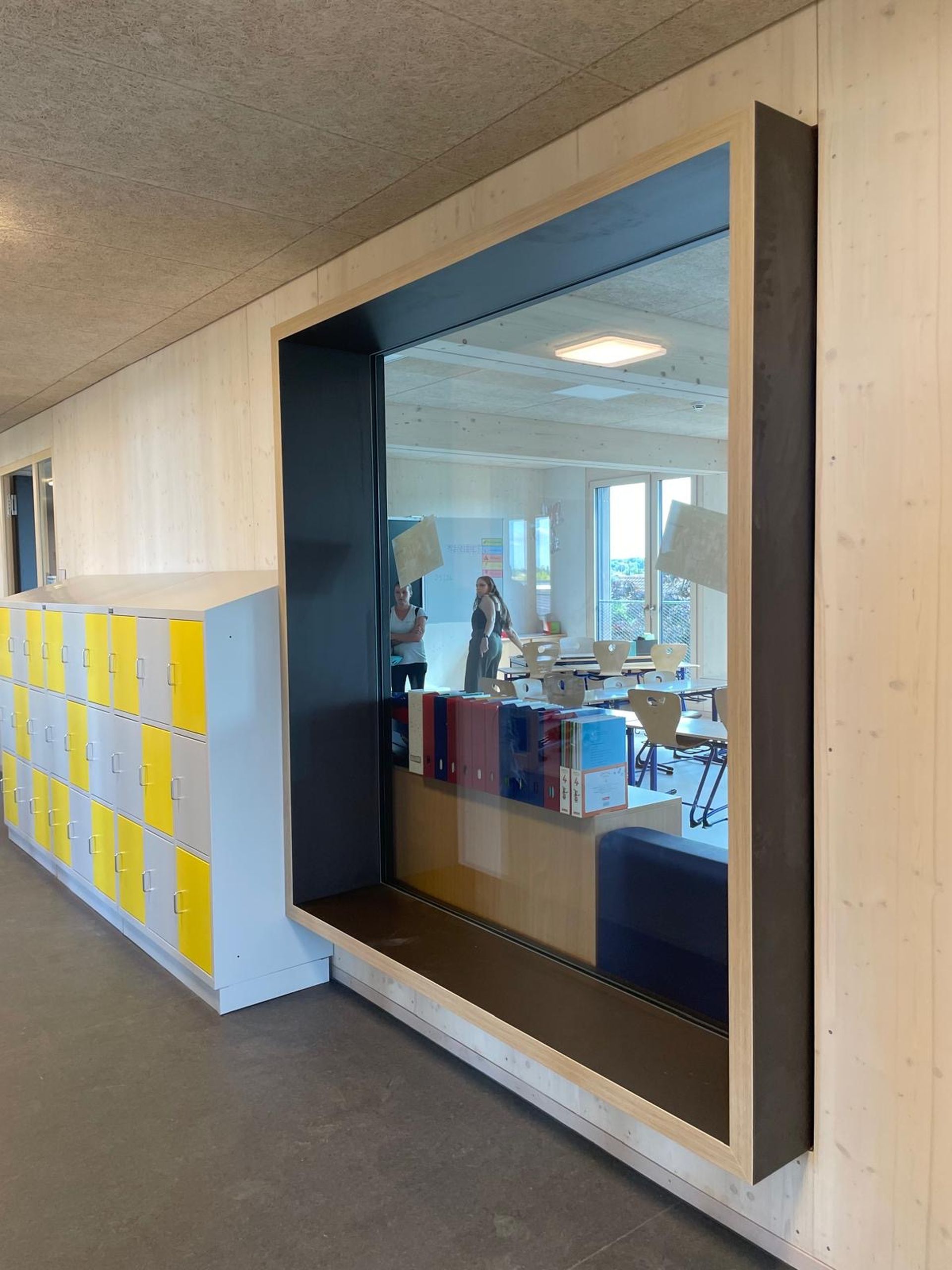Großes Fenster mit schwarzem Rahmen in einem modernen Flur, in dem sich Menschen und ein Klassenzimmer spiegeln. Gelbe Schließfächer säumen die Wand links.