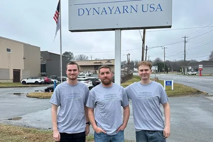 Drei Personen mit grauen T-Shirts mit der Aufschrift "Zimmermann- Dynayarn USA" stehen vor einem grauen Schild.
