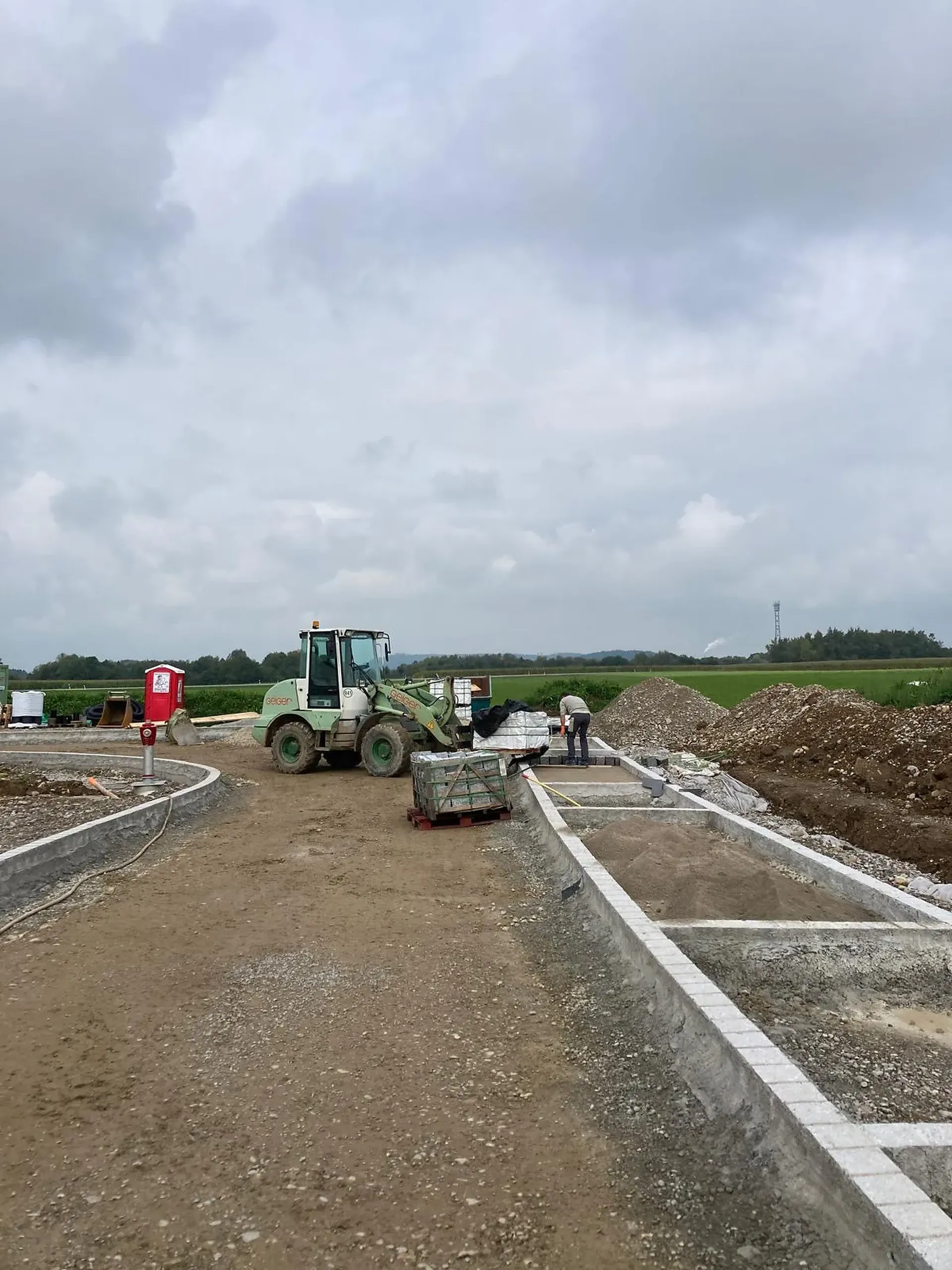 Baustelle mit einem Bulldozer, Arbeitern und Baumaterialien. Eine unbefestigte Straße und Betonstrukturen unter bewölktem Himmel in einer ländlichen Gegend.