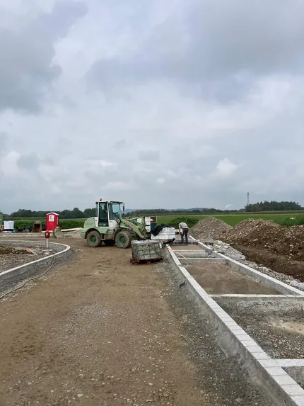 Baustelle mit einem Bulldozer, Arbeitern und Baumaterialien. Eine unbefestigte Straße und Betonstrukturen unter bewölktem Himmel in einer ländlichen Gegend.