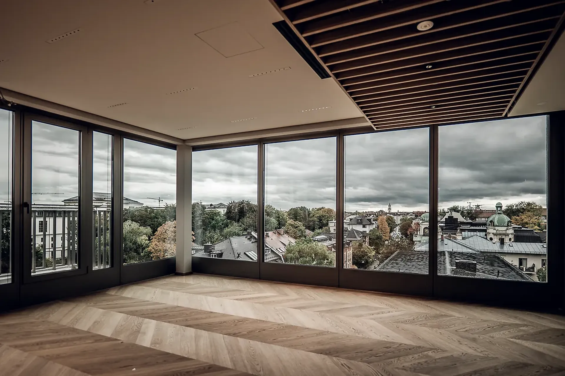 Geräumiges Zimmer im Bürohaus Novella 7 mit großen Fenstern und Blick auf die Stadtlandschaft unter einem bewölkten Himmel, mit Fischgrätholzboden und moderner Deckengestaltung.