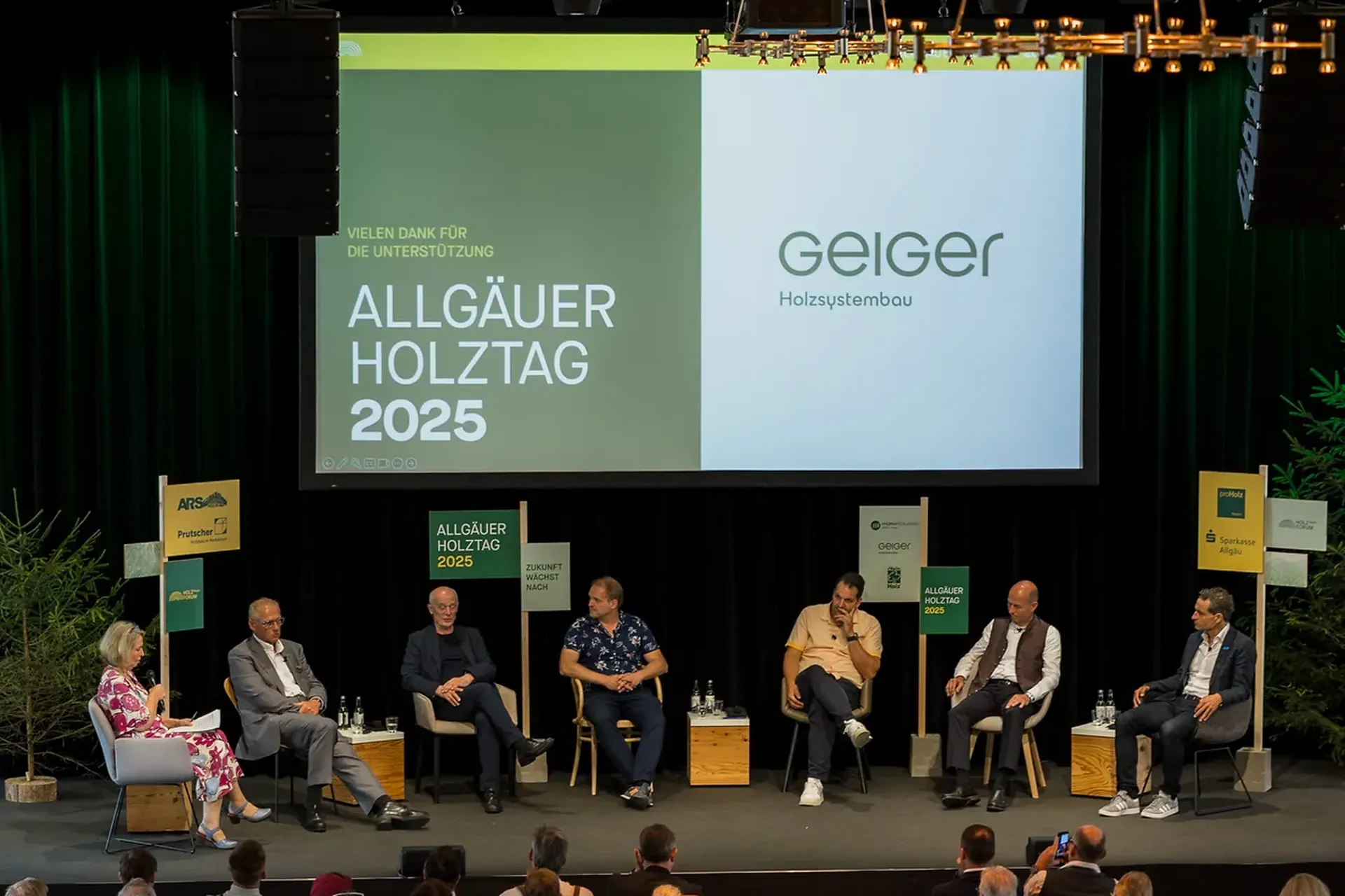 Podiumsdiskussion auf einer Bühne beim Allgäuer Holztag 2025 mit sechs Rednern, die unter einem modernen Kronleuchter sitzen und einem Publikum zugewandt sind.