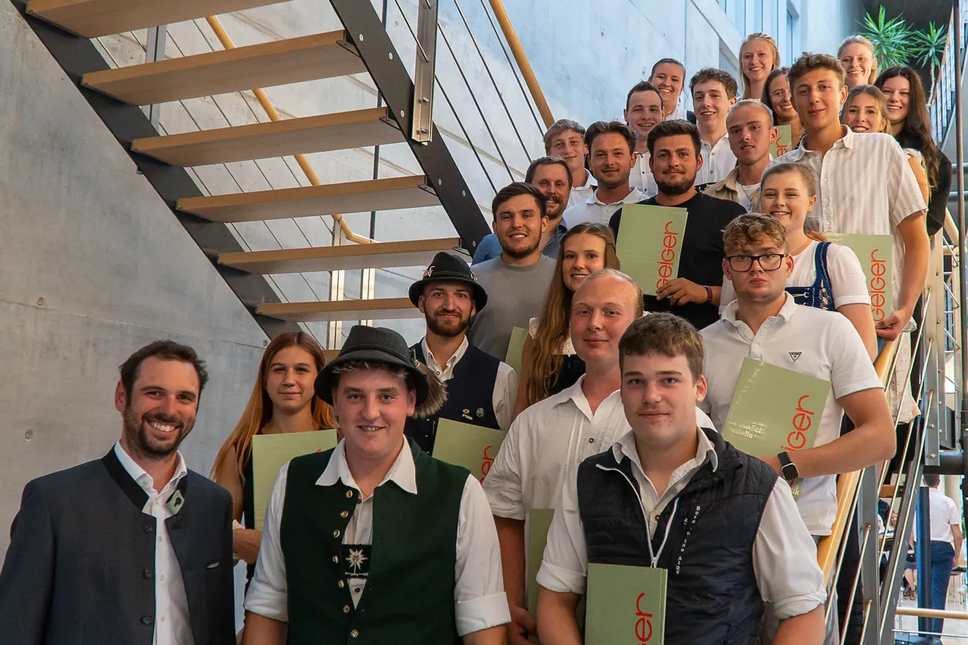 Die Gruppe der Auszubildenden steht drinnen auf einer Treppe, hält Zertifikate in den Händen und lächelt in die Kamera.